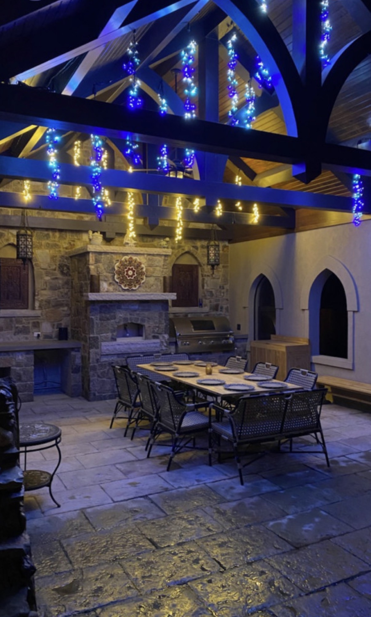 Illuminated timber pavilion interior with stone fireplace, outdoor kitchen and dining area.
